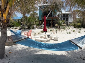 Un hamac, un parasol rouge et une chaise de plage, le tout installé sur une plage de sable, avec des palmiers et un bâtiment en arrière-plan.