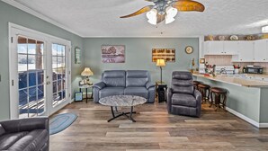Un salon spacieux doté d’un ventilateur de plafond, d’un canapé gris, d’une table basse et d’un coin cuisine avec un bar.