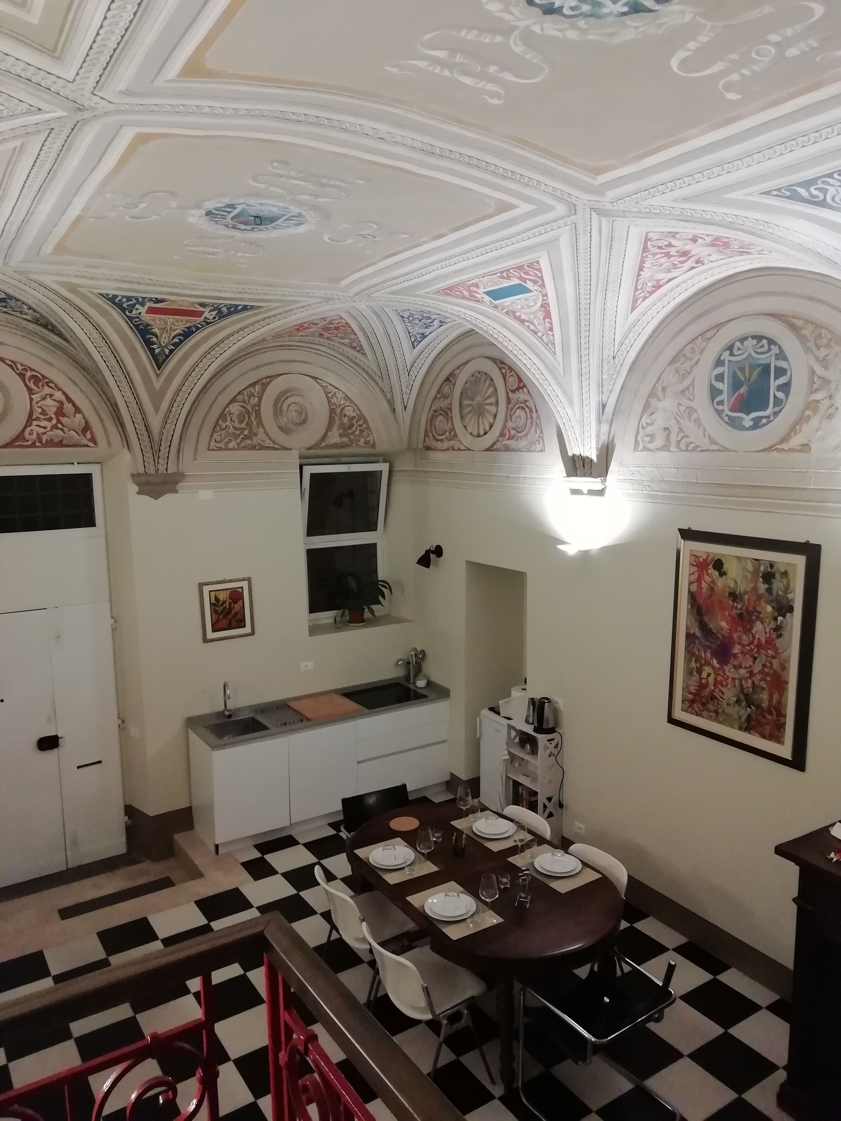 Ein Zimmer mit kariertem Boden, einem Esstisch für vier Personen, einer Küchenzeile mit Spüle und einer dekorativen Decke mit aufwendigen Verzierungen.