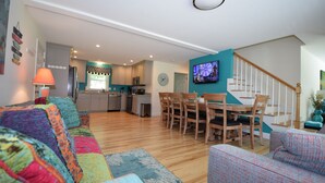 Un salon spacieux avec un ensemble de canapés aux couleurs vives, un coin repas avec des chaises en bois, une cuisine à l’arrière-plan et un escalier agrémenté d’un mur d’accent de couleur turquoise.
