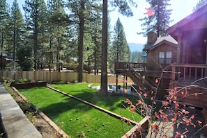 A wooden cabin with a deck, surrounded by tall trees and a well-maintained lawn.