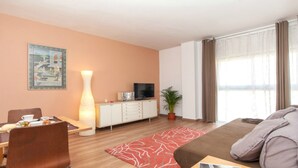 Una habitación de hotel moderna con zona de comedor, televisor, un florero grande y ventana con cortinas.