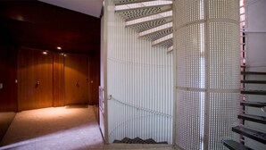 Un pasillo con una puerta de madera al final, una escalera en espiral blanca con barandilla de metal perforado y suelo alfombrado.