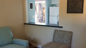 A living room with a sofa, a patterned armchair, a coffee table with a remote control, a black shelf, and a refrigerator in the background.