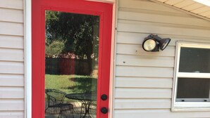A red door with a glass panel, a small outdoor table and chairs, and a wall-mounted light fixture.