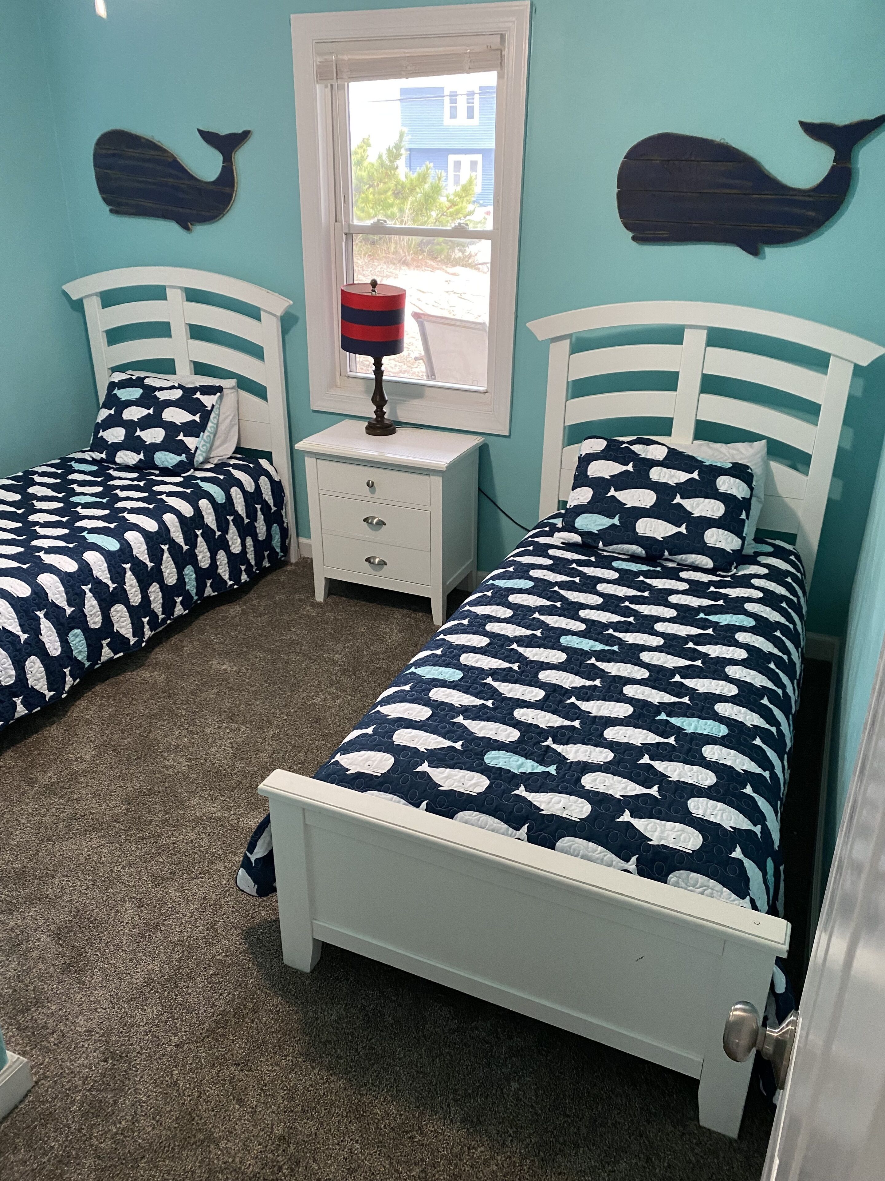 A bedroom with two beds, each with a white headboard and a blue and white fish-patterned bedspread. There is a nightstand with a lamp between the beds, and a window with a view of the outside.