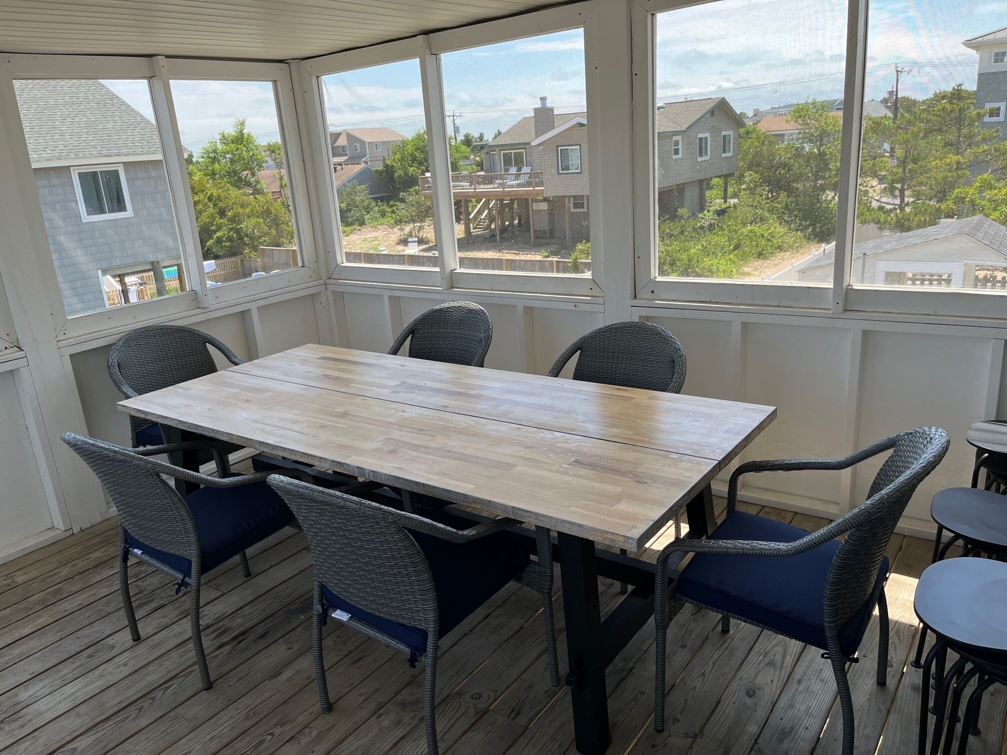 Une terrasse couverte avec une table en bois et des chaises en osier, de grandes fenêtres offrant une vue sur les maisons voisines.