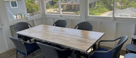 Une terrasse couverte avec une table en bois et des chaises en osier, de grandes fenêtres offrant une vue sur les maisons voisines.