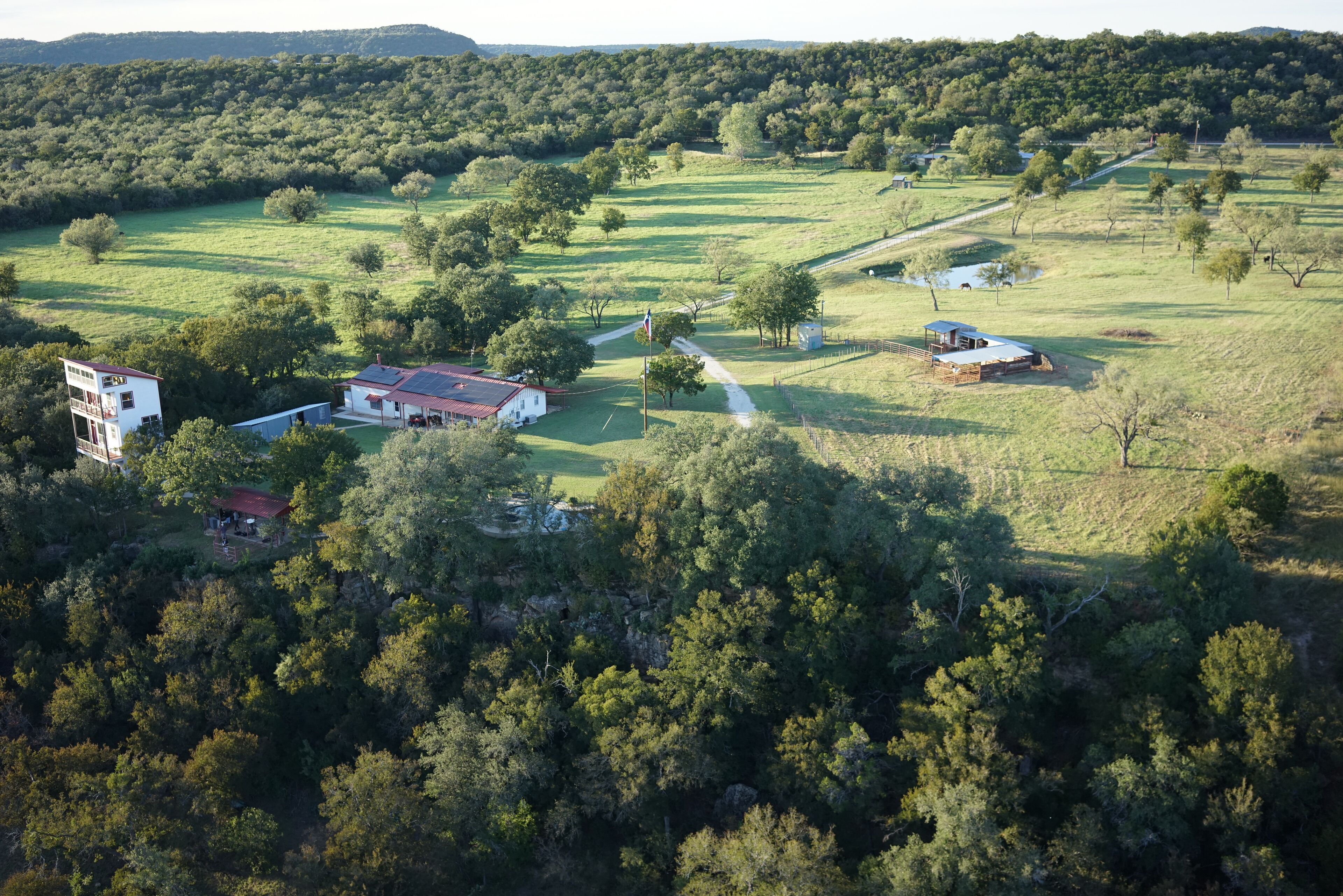 An aerial view of a rural property with multiple buildings, including a large white house with a red roof, a smaller house with a red roof, and a barn. There are several trees and a dirt path connecting the buildings.