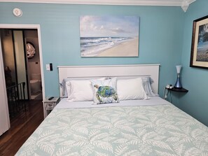 A bedroom with a bed, a painting of a beach, a nightstand, and a bathroom visible through an open door.