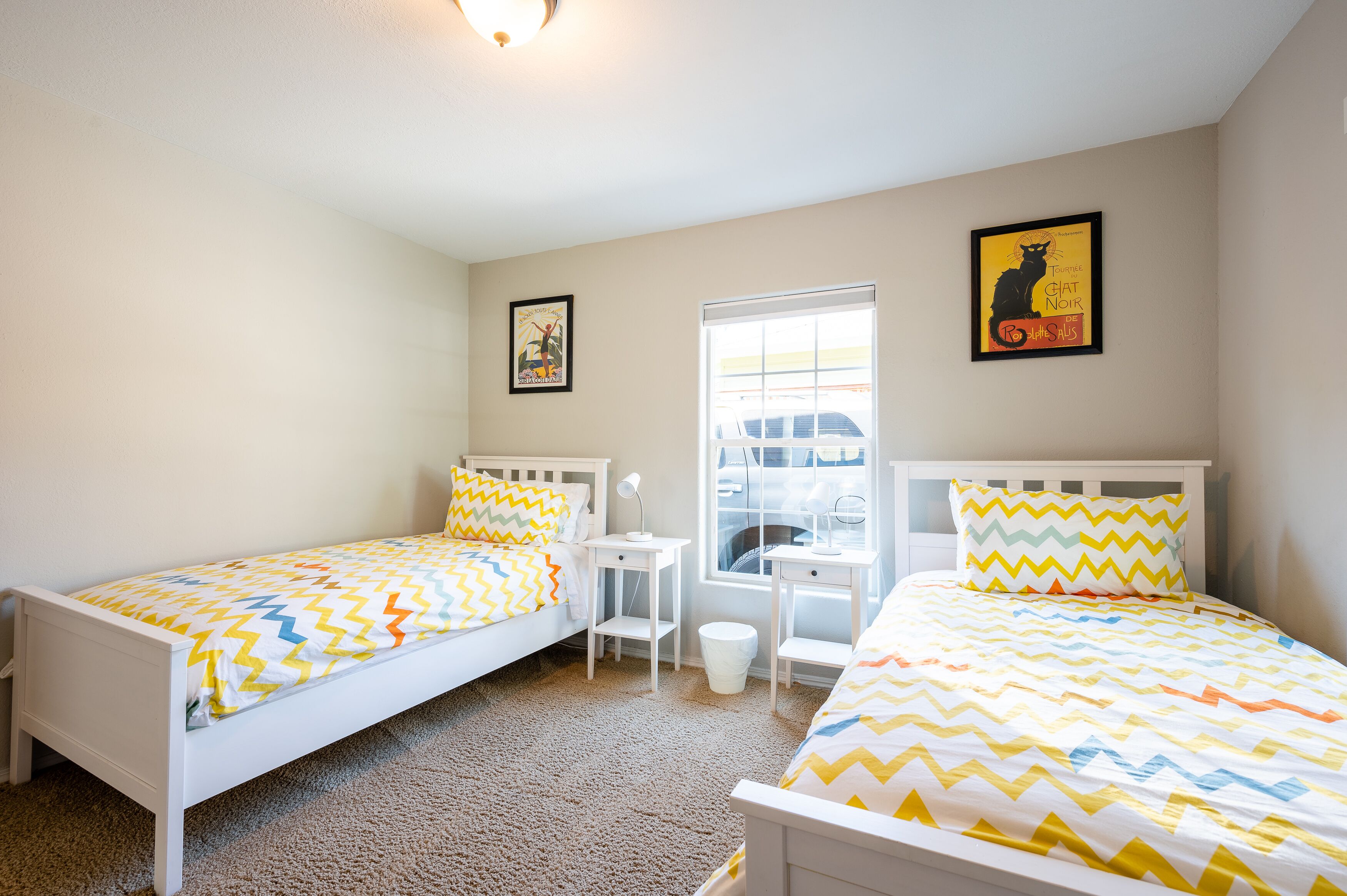 A bedroom with two single beds, a small white desk, and a window with a view of a car.