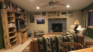Uma sala de estar aconchegante, com lareira, televisão e estante repleta de livros e objetos decorativos.
