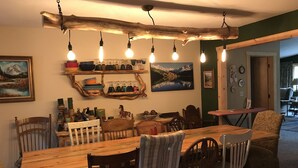 Uma área de jantar com uma longa mesa de madeira, várias cadeiras, lâmpadas pendentes e prateleiras com diversos objetos.