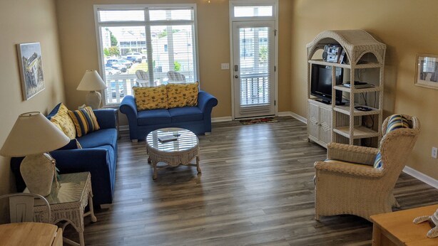A living room with a blue sofa, a round coffee table, wicker chairs, and a TV stand. - On Sunset Beach Island 1 Bedroom Condo sleeps 4 Water view! Unit 2 (Sunset Beach)
