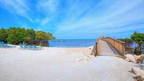 A beach with a wooden walkway leading to a gazebo, surrounded by trees and clear skies.