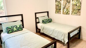 Een kamer met twee eenpersoonsbedden, elk met een houten frame en licht beddengoed. Drie kunstwerken aan de muur met afbeeldingen van tropische bladeren.
