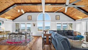 Een ruime woonkamer met een hoge plafond, houten balken, een groot raam met uitzicht op zee, een grijze bank en een eethoek met een tafel en stoelen.
