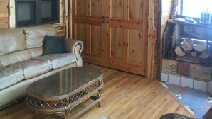 Een gezellig interieur van een blokhut met een houten plafond, een grote kledingkast, een comfortabele bank, een rieten tafeltje en een open haard met hout.