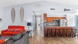 A modern living room with a red sofa, a grey couch, and a small table with a lamp. A kitchen area with wooden cabinets, a microwave, and a bar stool is visible.