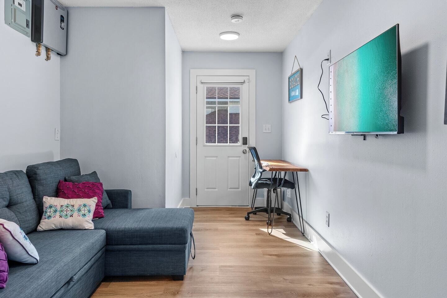 Ein modernes Wohnzimmer mit einem grauen Ecksofa, einem Holzesstisch und einem schwarzen Bürostuhl. Im Hintergrund ist eine weiße Tür mit Fenster erkennbar.