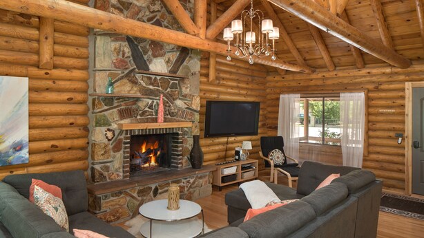 A log cabin interior with a stone fireplace, a television mounted on the wall, and a large gray sectional sofa.
