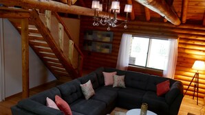 A cozy log cabin interior with a sectional sofa, a chandelier, a staircase, and a large window.