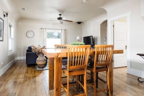 A dining area with a wooden table and chairs, a ceiling fan, a clock on the wall, and a window with curtains.