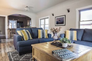 Een moderne woonkamer met een donkerblauwe zithoek, een houten salontafel en een groot raam dat voor natuurlijk licht zorgt.