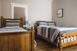 A bedroom with two wooden beds, a window with blinds, a bedside table with a lamp, and a framed picture on the wall.