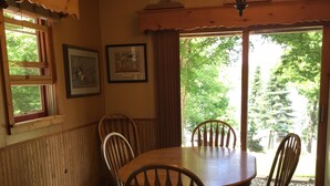 A dining area with a round wooden table and four chairs, a chandelier, framed pictures on the wall, and a window with a view of trees. - Summer is coming!   (Longville)