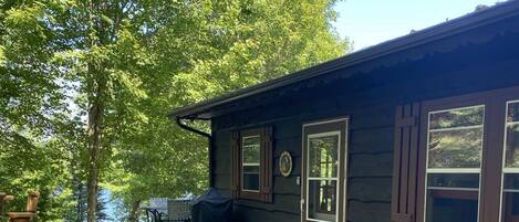 A cabin with a covered outdoor seating area, a covered trash bin, and a paved driveway with scattered leaves.