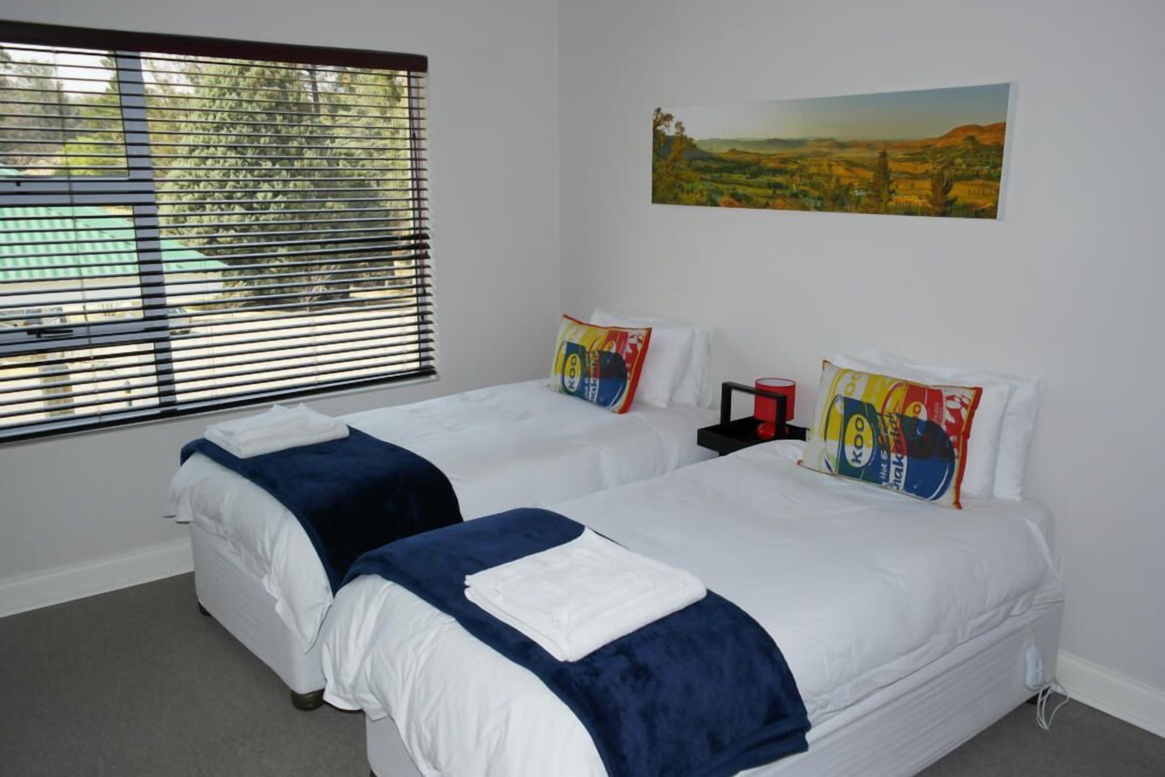 A hotel room with two beds, a window with blinds, a painting on the wall, and a red object on the nightstand.