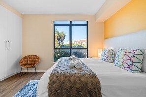 Une chambre d’hôtel avec une grande fenêtre, un lit avec du linge de lit blanc, un plaid à motifs et une chaise.