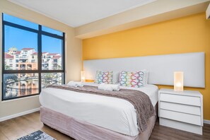A bedroom with a large window, a bed with white linens, a headboard, and bedside lamps. - Capri 101 (Cape Town)