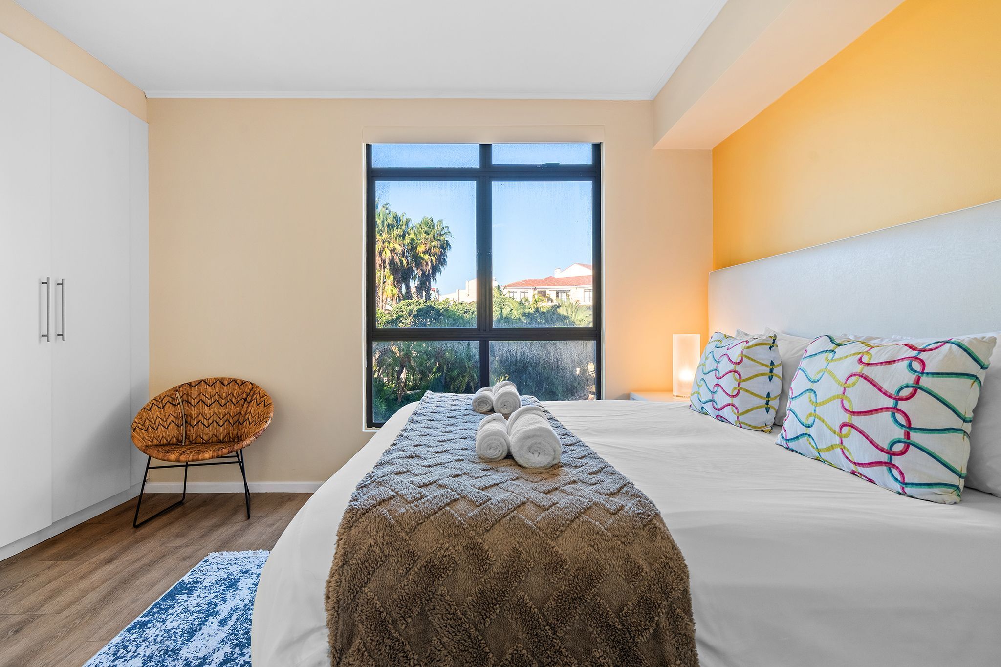 A bedroom with a large window, a bed with white linens, a patterned throw blanket, and a chair.