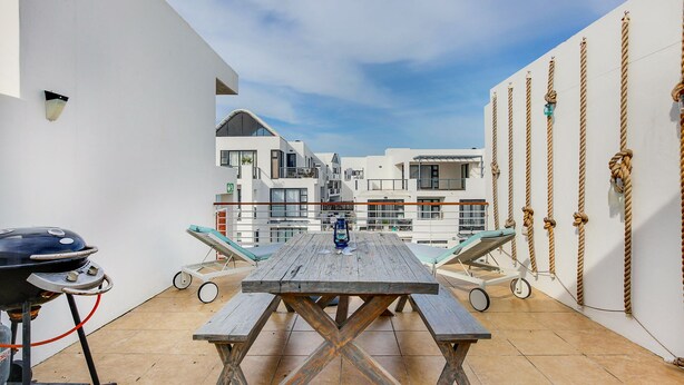 Una terraza en la azotea con mesa y bancos de madera, una parrilla y vistas a edificios blancos.