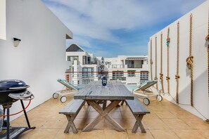 A rooftop patio with a wooden table and benches, a barbecue grill, and a view of white buildings. - Azure 208 On Big Bay (Cape Town)