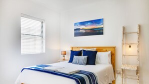 Un dormitorio con una cama de madera, ropa de cama blanca, una manta azul y blanca a rayas, un cuadro de un atardecer y una escalera de madera apoyada contra la pared.