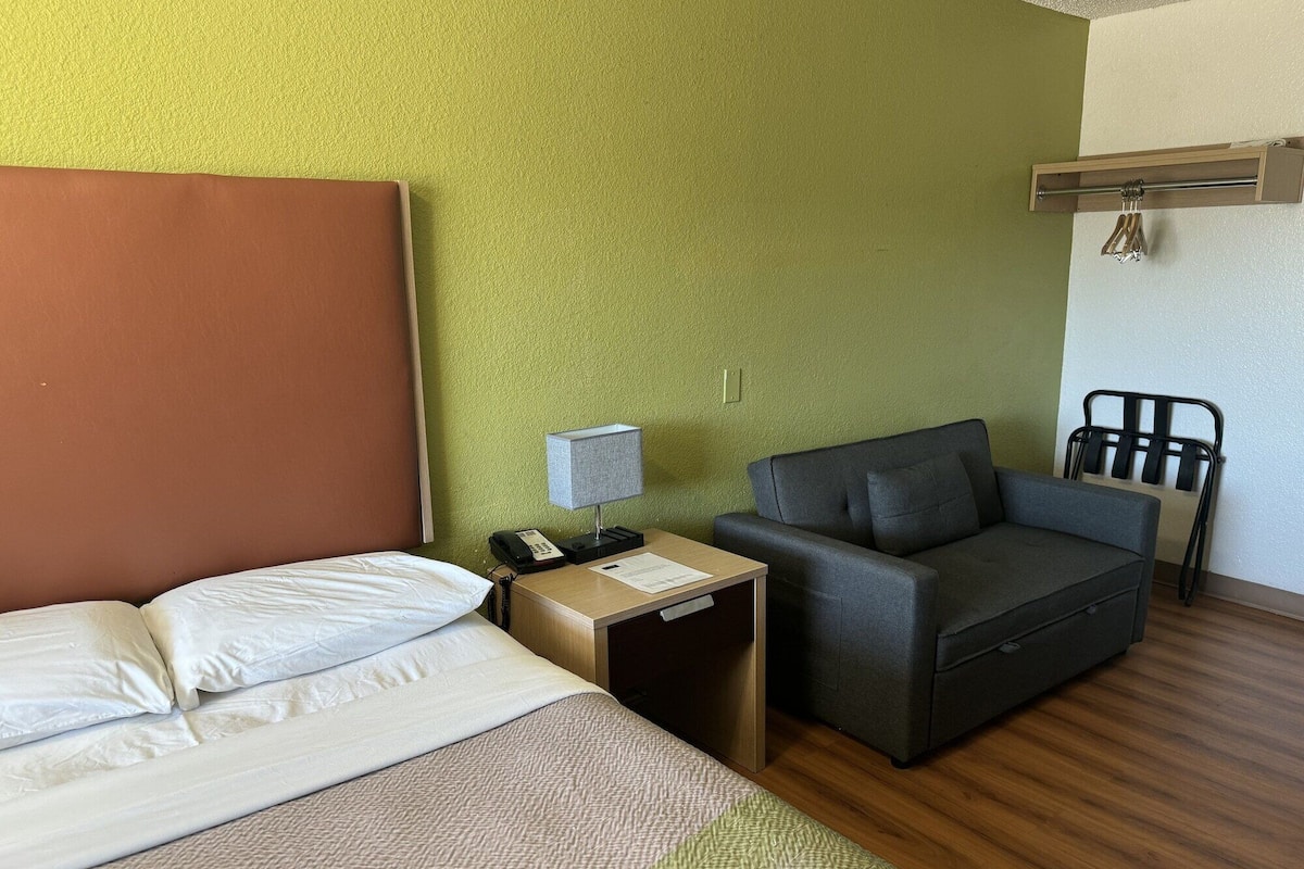 A hotel room with a bed, a desk with a lamp, a grey sofa, a chair, and a wooden headboard.