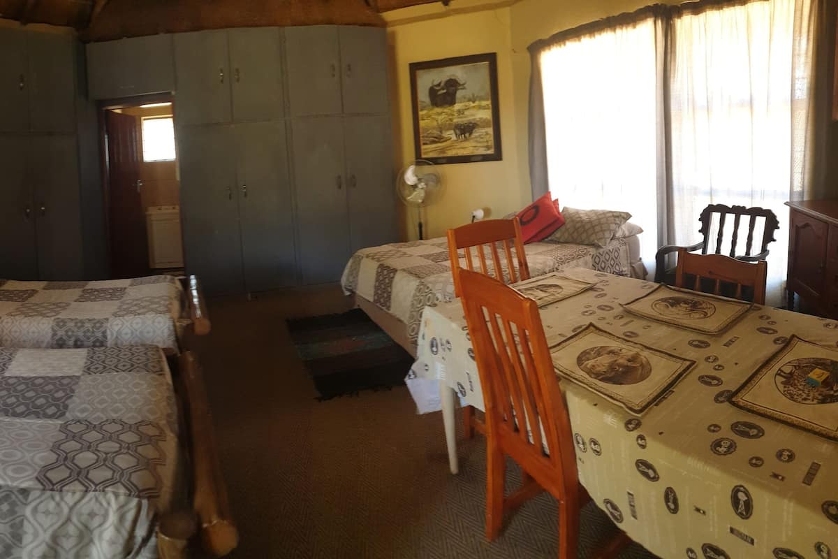 A room with two beds, a dining table, and a chair. There is a wardrobe and a painting on the wall.