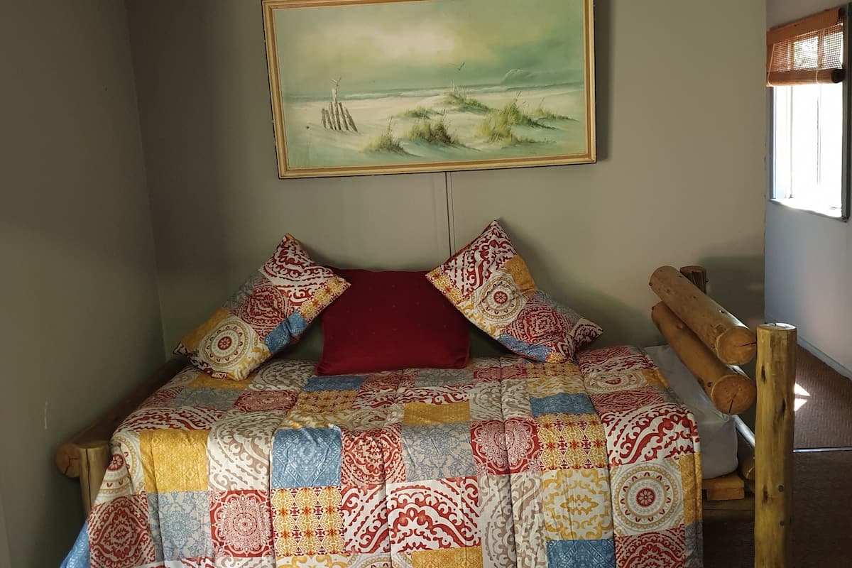 A bed with a colorful quilt, a red pillow, and a painting of a beach scene on the wall.