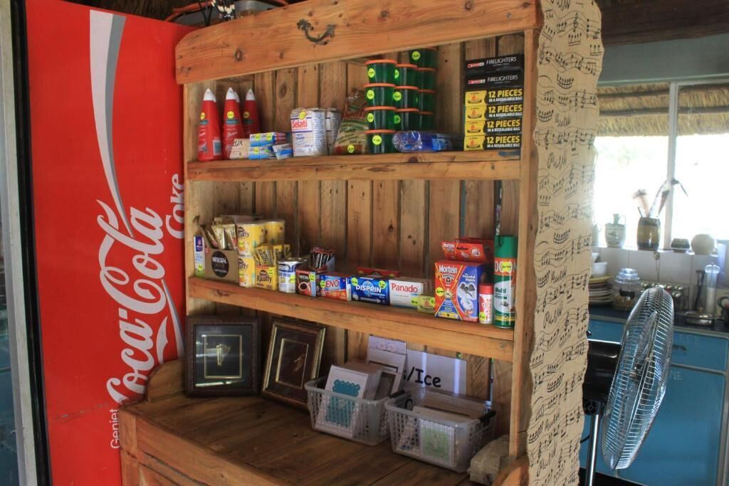 Une étagère en bois remplie de divers produits alimentaires et condiments, une enseigne Coca-Cola et un ventilateur à l’arrière-plan.