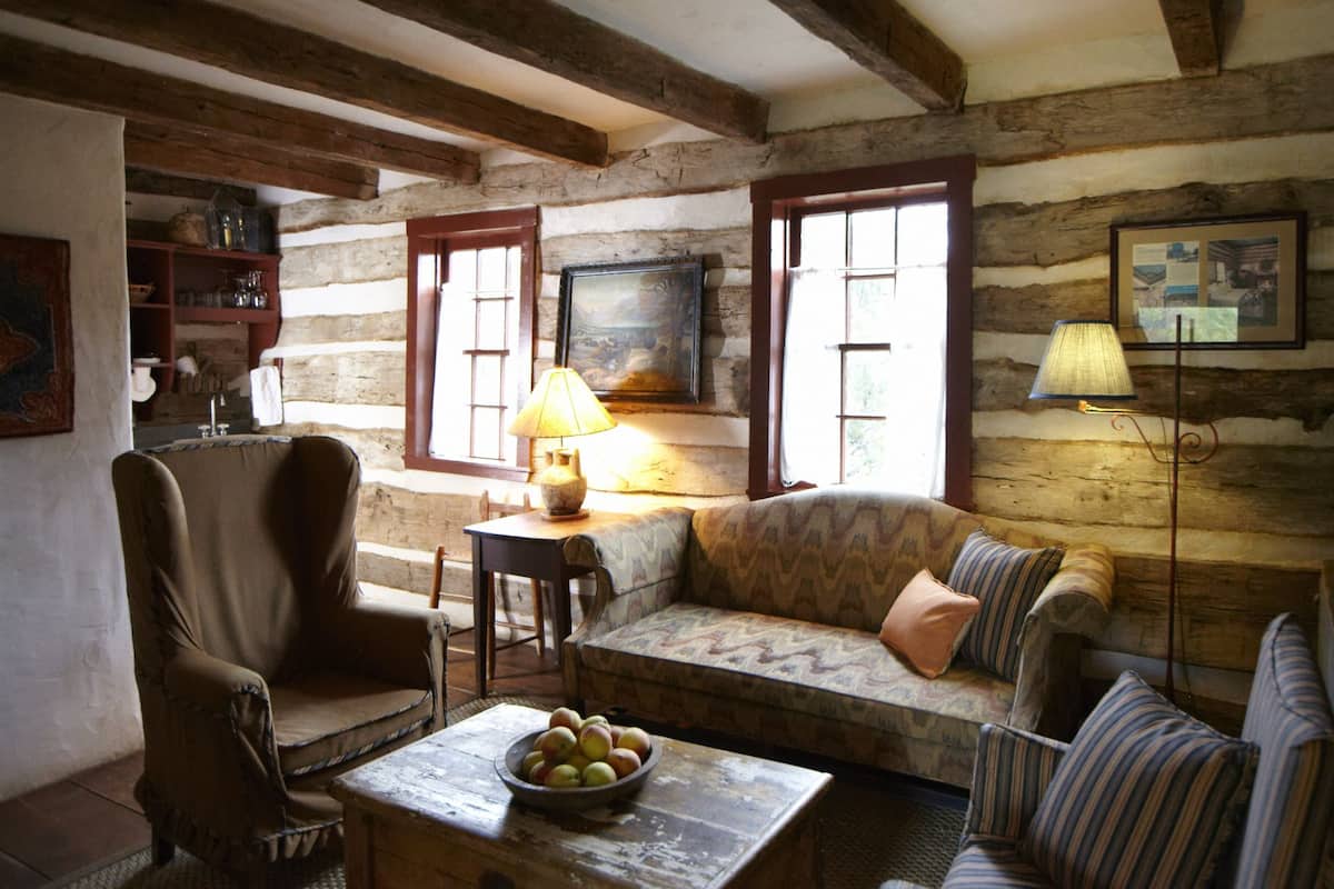 A rustic room with a wooden log wall, a sofa, a chair, a table with a bowl of fruit, and a lamp.