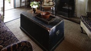 A rustic living room with a fireplace, wooden beams, a cabinet with dishes, and a patterned rug. - Settlers Crossing (Fredericksburg)