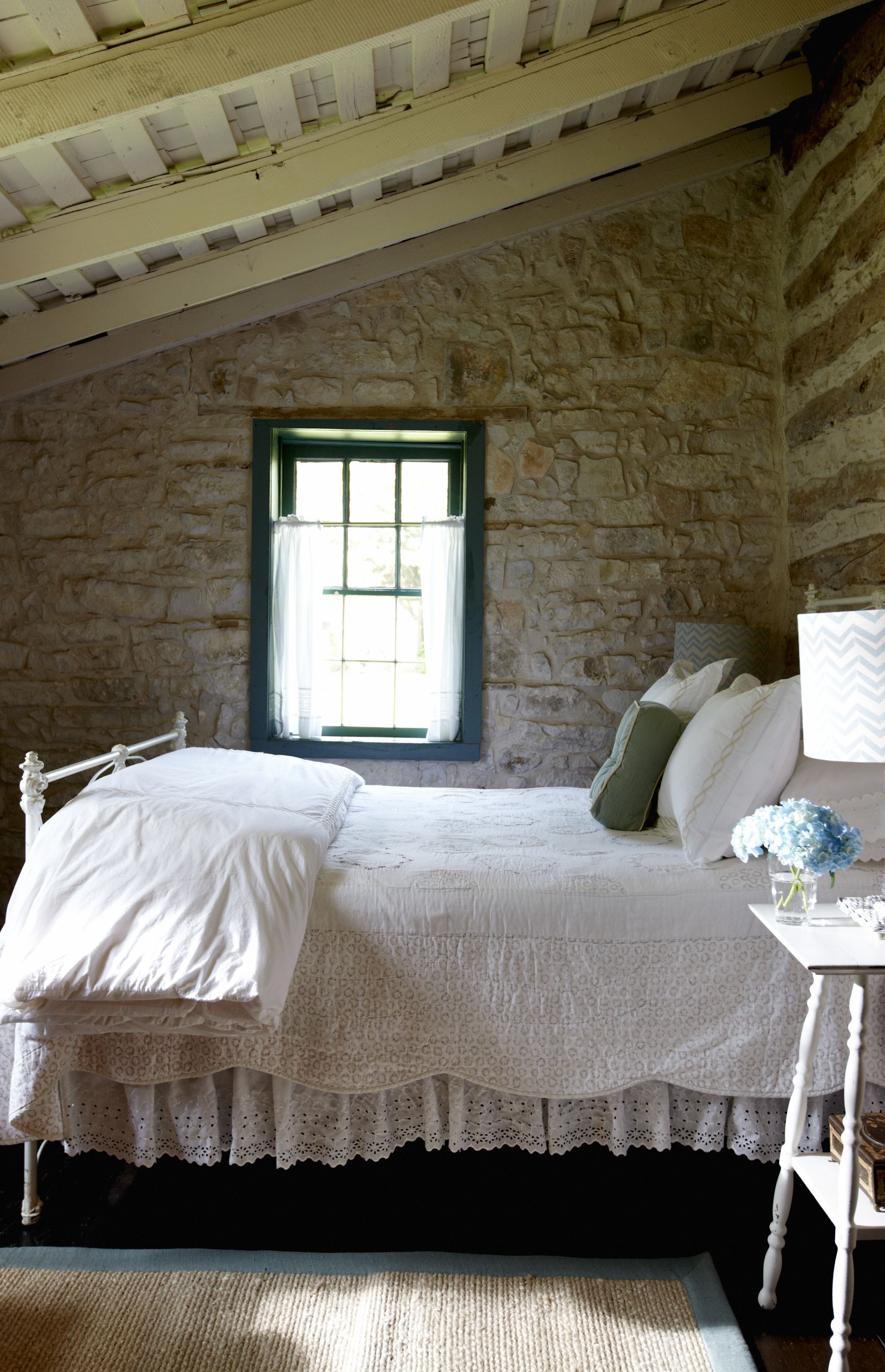 Une chambre avec un mur en pierre, un lit en bois avec une literie blanche, une fenêtre avec des volets bleus et une petite table sur laquelle est posé un vase de fleurs.