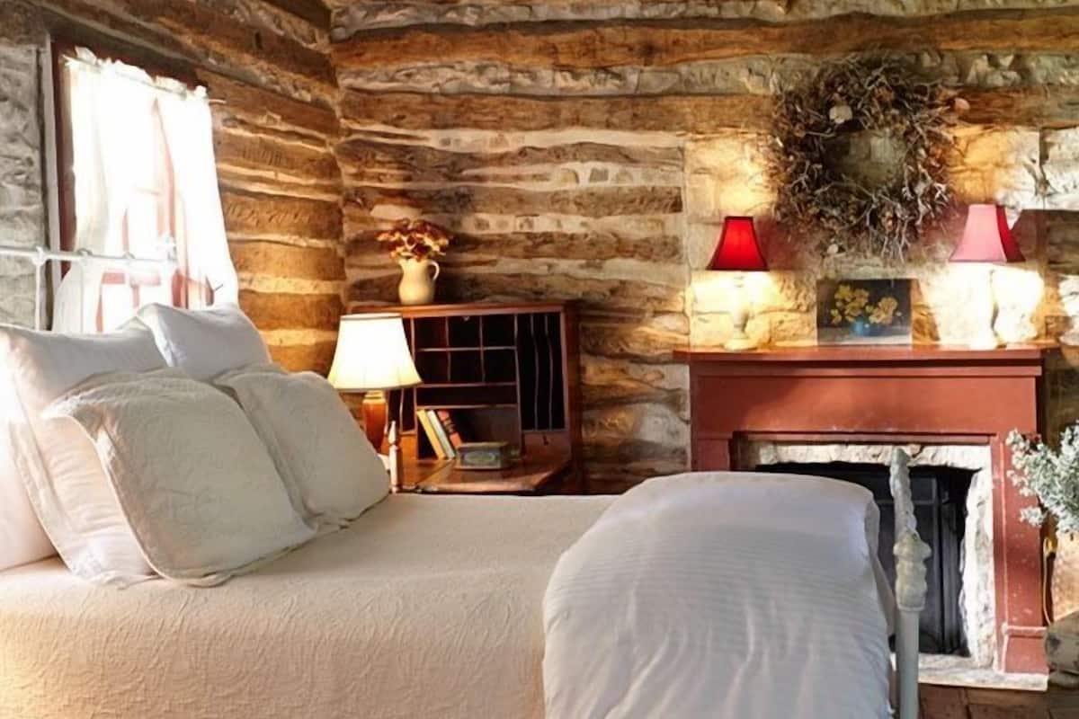 A rustic log cabin bedroom with a bed, a nightstand, a fireplace, and a window with curtains.