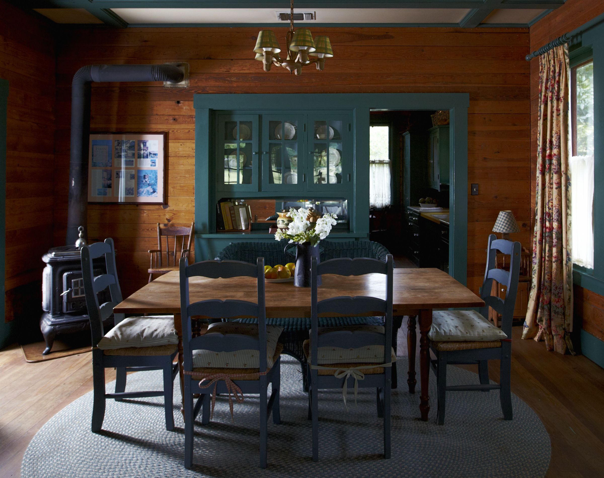 Une salle à manger avec une table en bois, six chaises, un vase de fleurs et une cuisine visible à travers une porte.