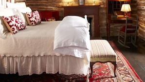 A log cabin-style bedroom with a bed, pillows, a bench, a fireplace, and two lamps. - Settlers Crossing (Fredericksburg)