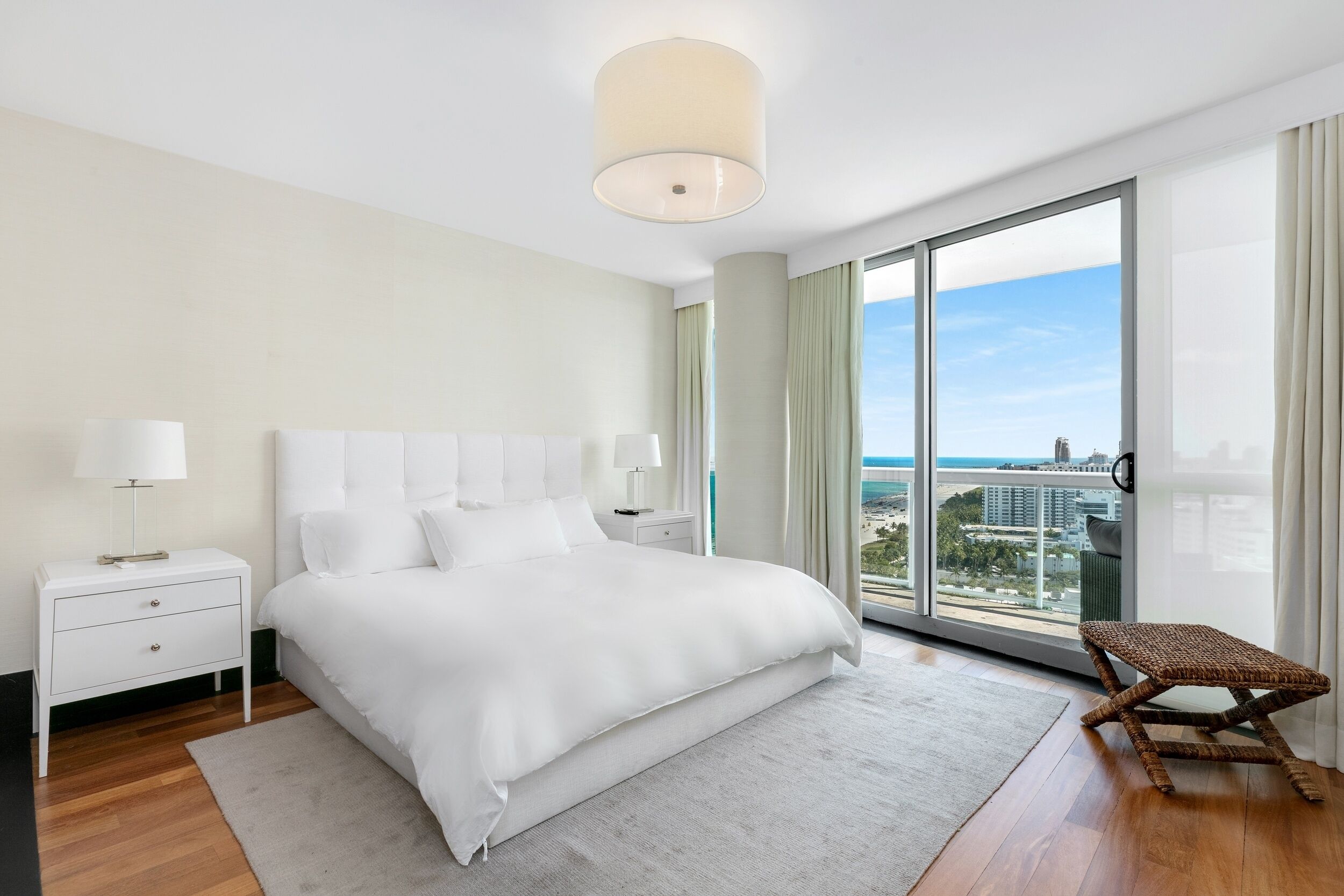 A modern hotel room with a large bed, a nightstand, a lamp, and a view of the city through large windows.