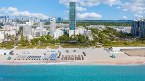 Un paysage urbain côtier avec un grand immeuble imposant, une plage avec des chaises longues et une vue dégagée sur l’océan.